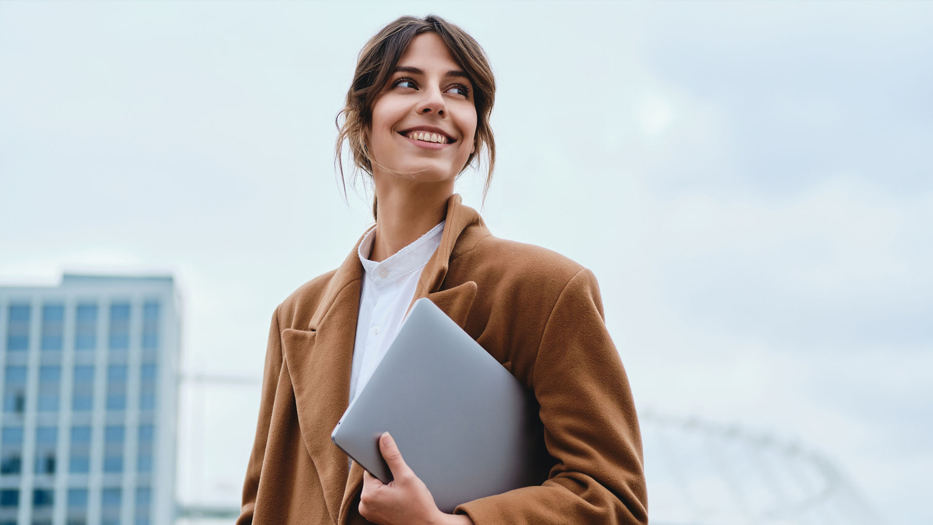 Young pretty smiling businesswoman in coat with laptop happily looking away on city street