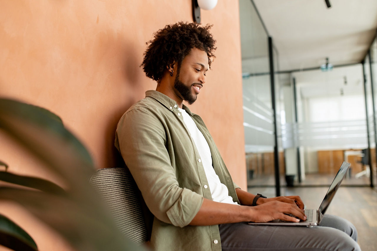 African-American male employee, freelance, programmer typing on laptop in office hallway, positive and smiling team member responding emails, chatting online with colleagues