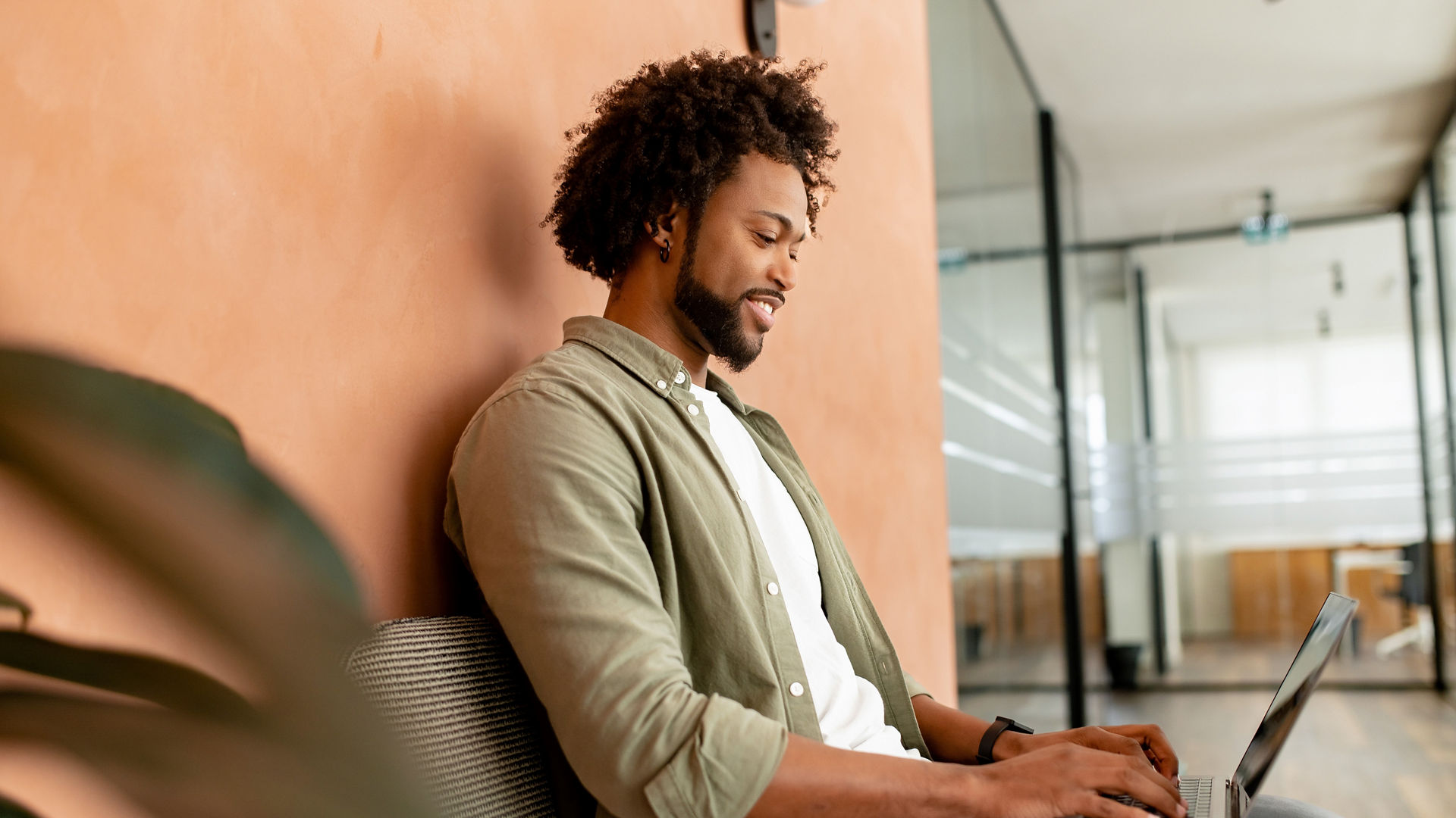 African-American male employee, freelance, programmer typing on laptop in office hallway, positive and smiling team member responding emails, chatting online with colleagues