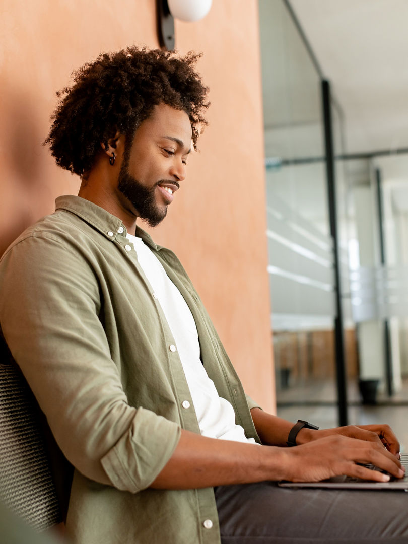 African-American male employee, freelance, programmer typing on laptop in office hallway, positive and smiling team member responding emails, chatting online with colleagues