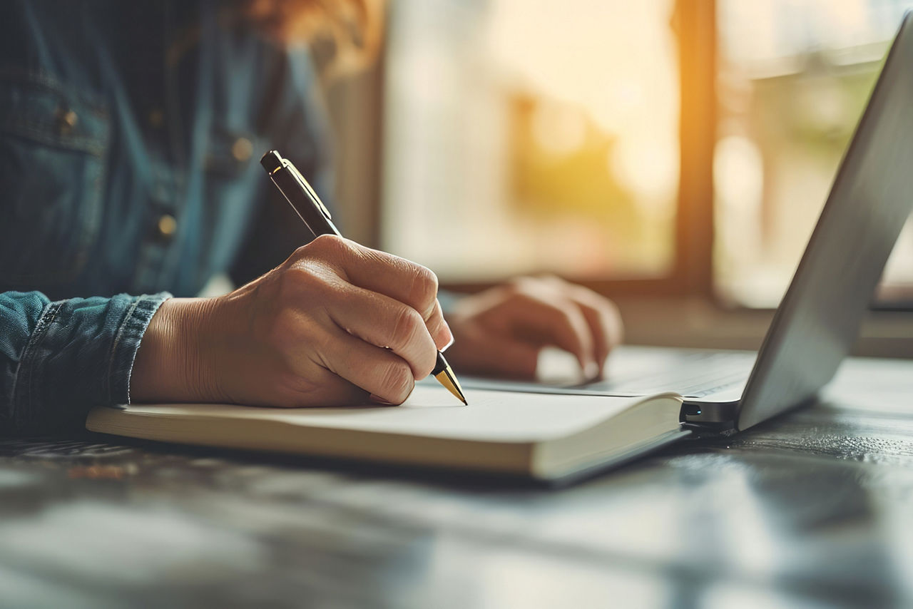 Close up of a person hands writing in a notebook with a pen, with a laptop in the background.