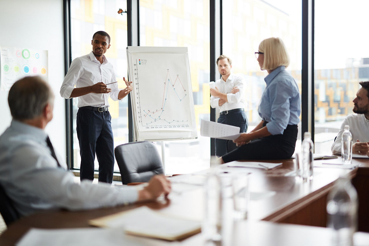 One of young businessmen explaining financial data to colleagues while standing by whiteboard