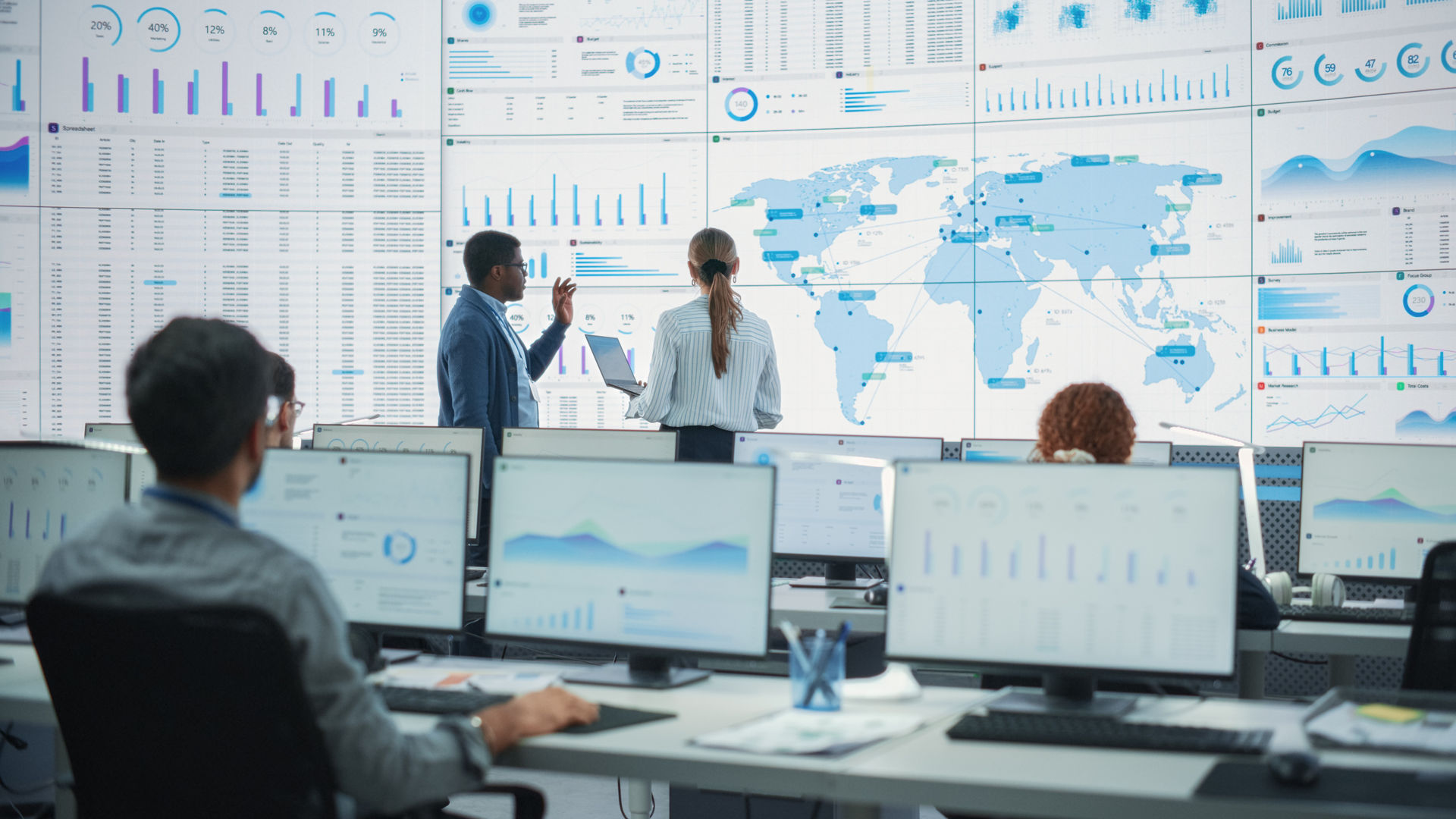 Back View Of Black Male Analyst Talking To Caucasian Female Manager In Front Of Big Digital Screen With World Map In Monitoring Office. Diverse Employees Working Behind Computers In Logistics Company.